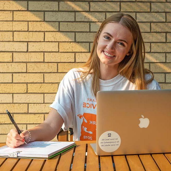 Smiling student outside with laptop and notebook on table in front of her