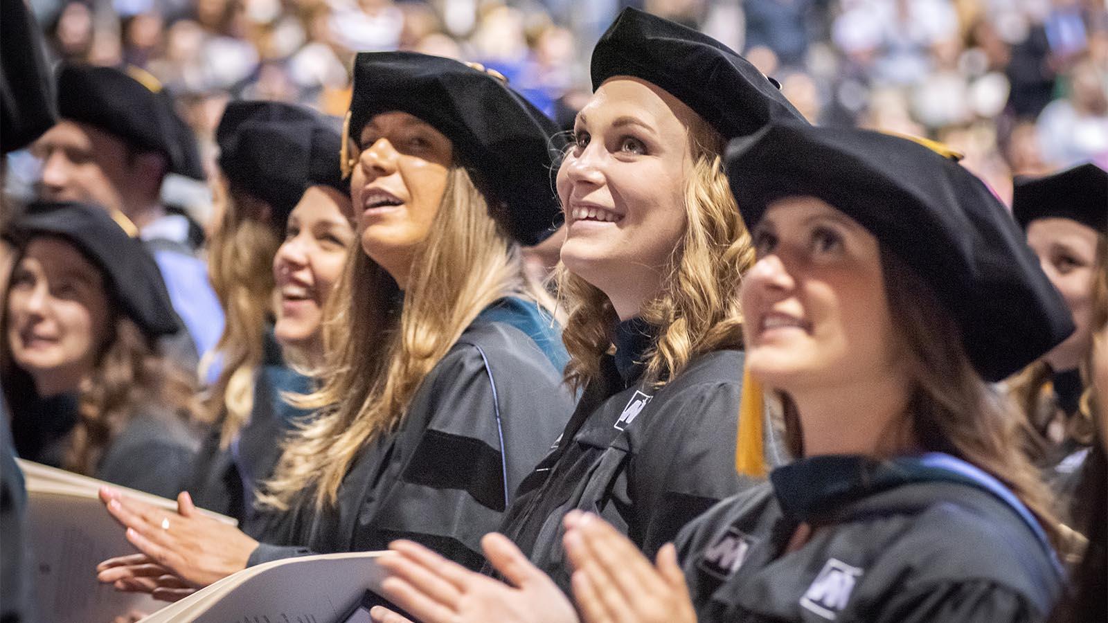 Three female University of Mary students sitting among a crowd of their fellow graduates.